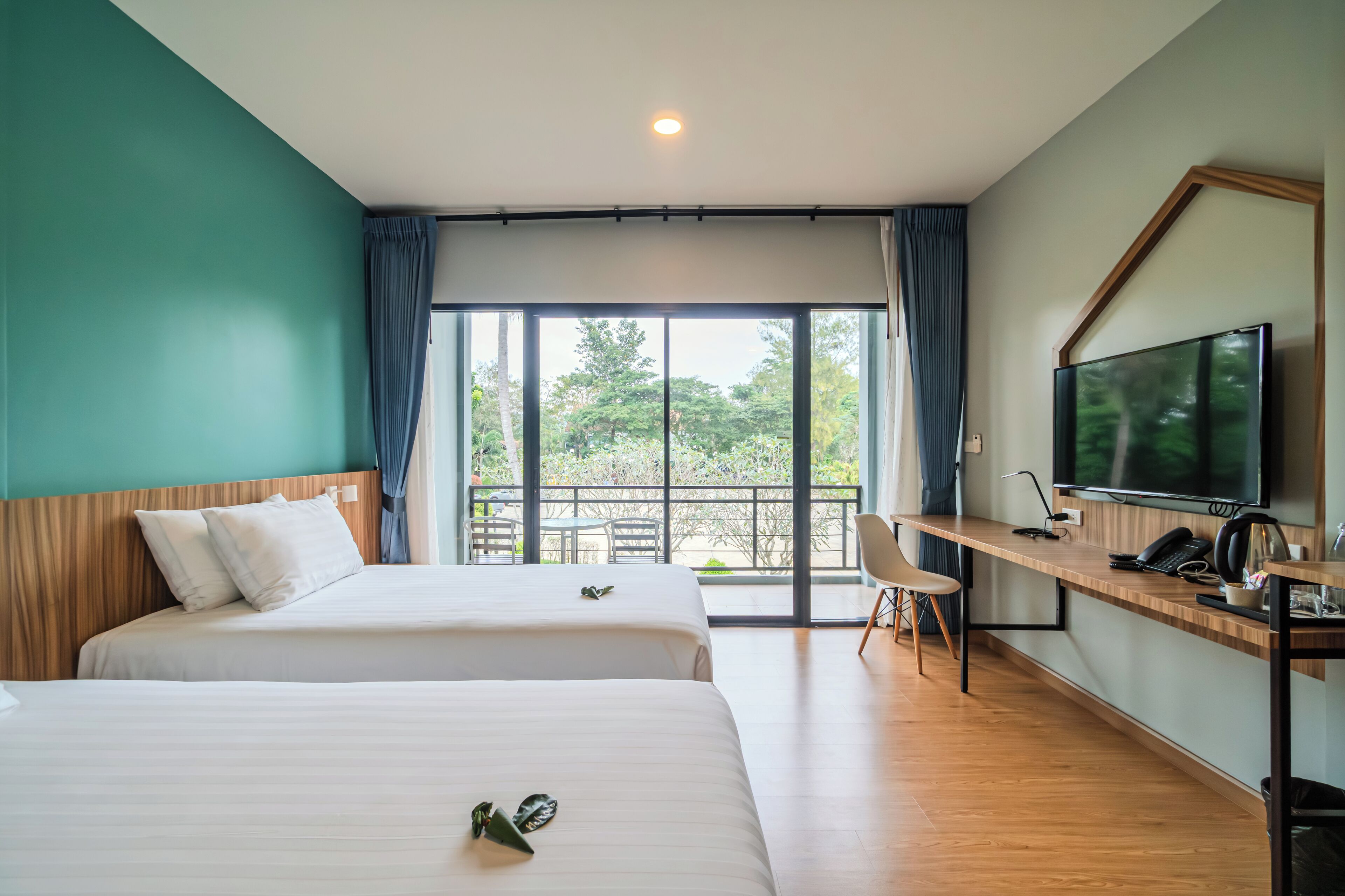Habitación de hotel con dos camas, un escritorio con computadora, televisor y balcón con vista a árboles.