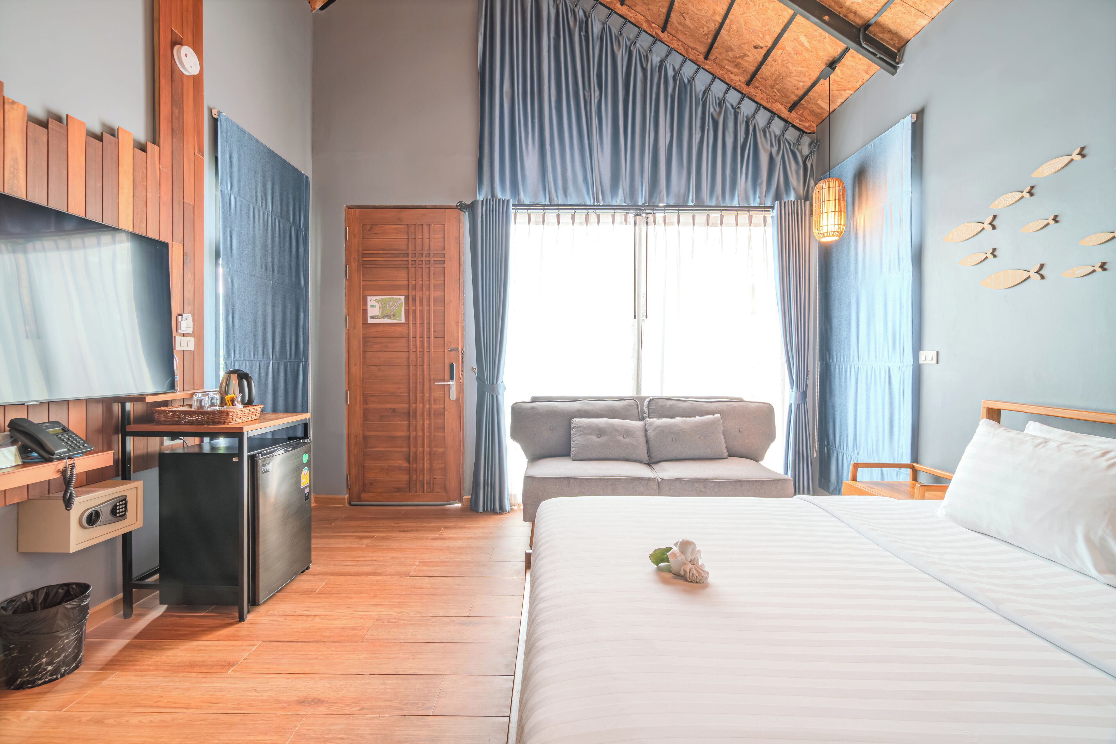 Una camera d'hotel con un letto, una scrivania con frigorifero, un armadietto in legno, una finestra con tende e una parete decorata con pesci.