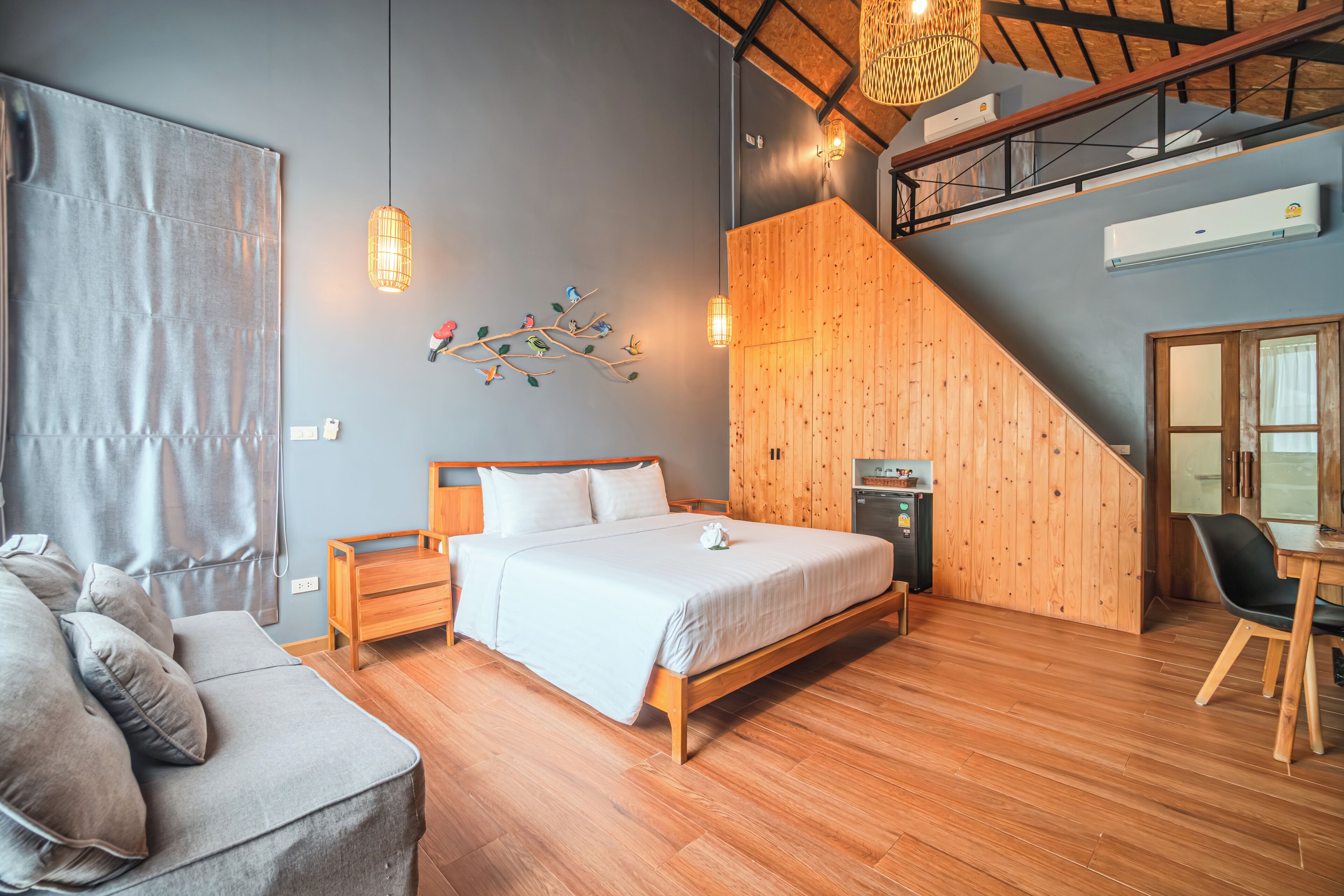 A bedroom with a wooden bed, a grey sofa, a wooden desk, and a staircase.