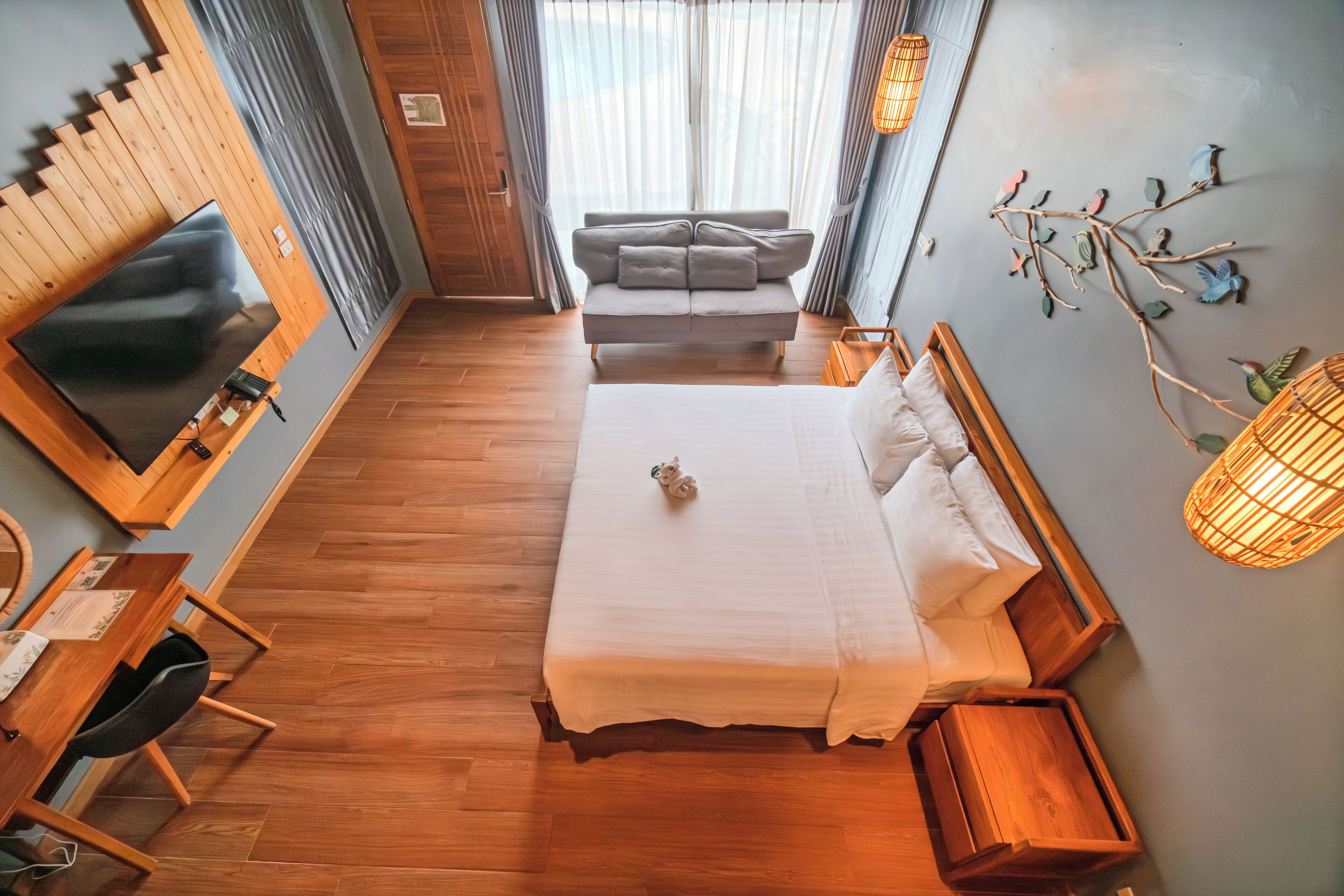 Ein modernes Hotelzimmer mit Holzboden, einem großen Bett mit weißer Bettwäsche, einem grauen Sofa, einem flachbildfernseher, der an einer Holzvertäfelung angebracht ist, und einer Wandleuchte.