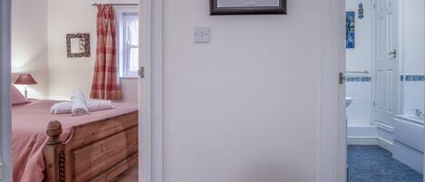 A bedroom with a wooden bed, a bedside table, and a lamp. There is a bathroom visible with a sink and a mirror.