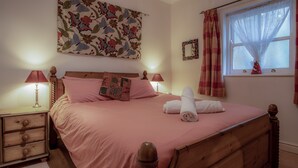 Quarto com cama de madeira, roupa de cama rosa, duas luminárias de apoio, tapeçaria floral, janela com cortinas e um pequeno espelho na parede.