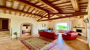 Un'ampia zona giorno con soffitto in legno a vista, un camino e una grande finestra con vista sul verde.
