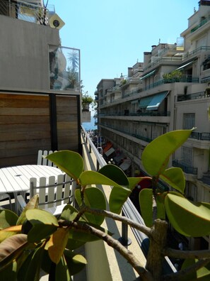 A balcony with a view of a coastal street, featuring a wooden table and chairs, and a plant with broad leaves. - Beautiful loft studio in Nice center, close to beach ! (Nice)