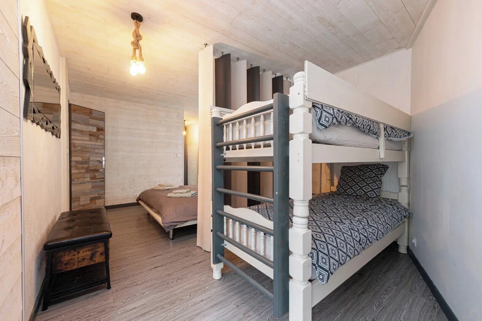 A bunk bed room with a wooden floor, a small bedside table, and a wall-mounted light fixture.