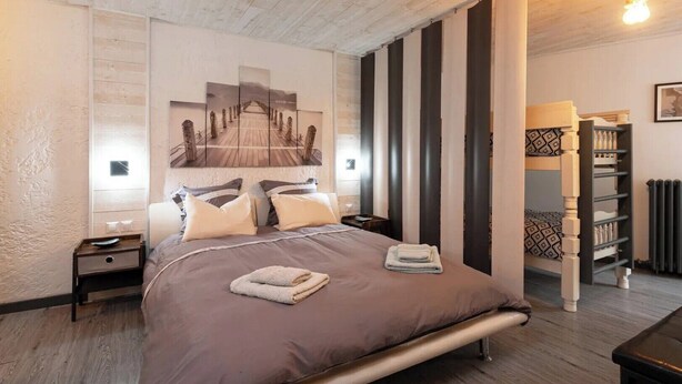 Une chambre à coucher avec un lit, des oreillers, une table de chevet, une œuvre d’art accrochée au mur, un radiateur et un plancher en bois.