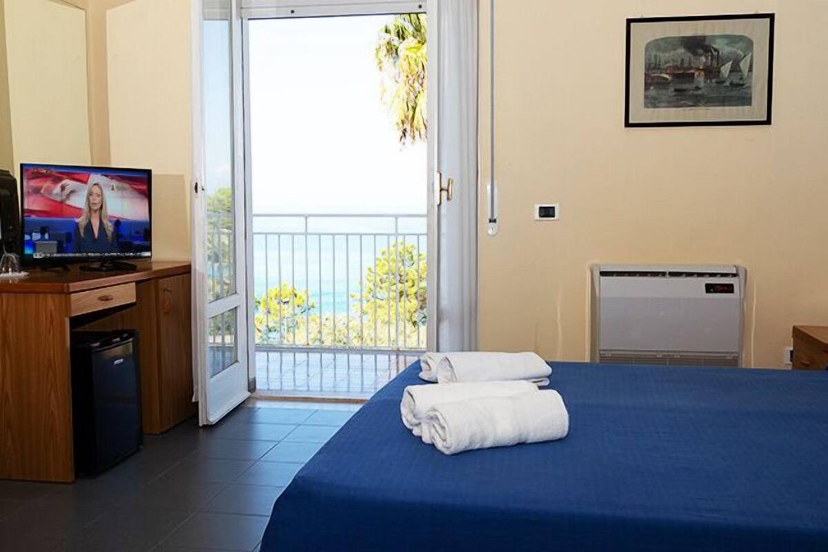 Ein Hotelzimmer mit Bett, Fernseher, Schreibtisch mit Computer, kleinem Kühlschrank, Heizkörper und einem Balkon mit Meerblick.