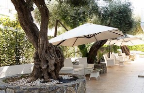 Un área de estar al aire libre con sillas blancas y sombrillas grandes, rodeada de árboles y vegetación.