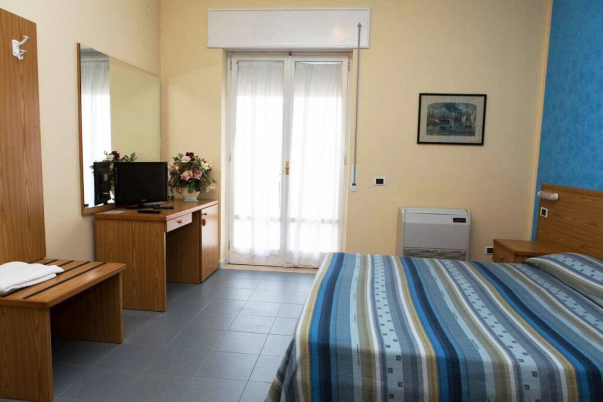 Ein Hotelzimmer mit einem Bett, einem Schreibtisch mit Computer, einem Stuhl, einer Bank, einem Fenster mit Vorhängen und einer Wand-Klimaanlage.