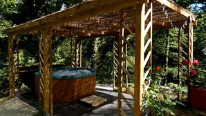 A wooden gazebo with a hot tub, surrounded by lush greenery and red flowers.