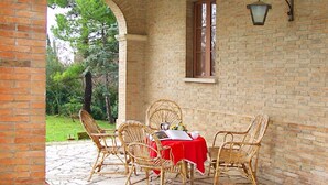 Une terrasse extérieure avec un mur en briques, une table recouverte d’une nappe rouge et des chaises en osier.