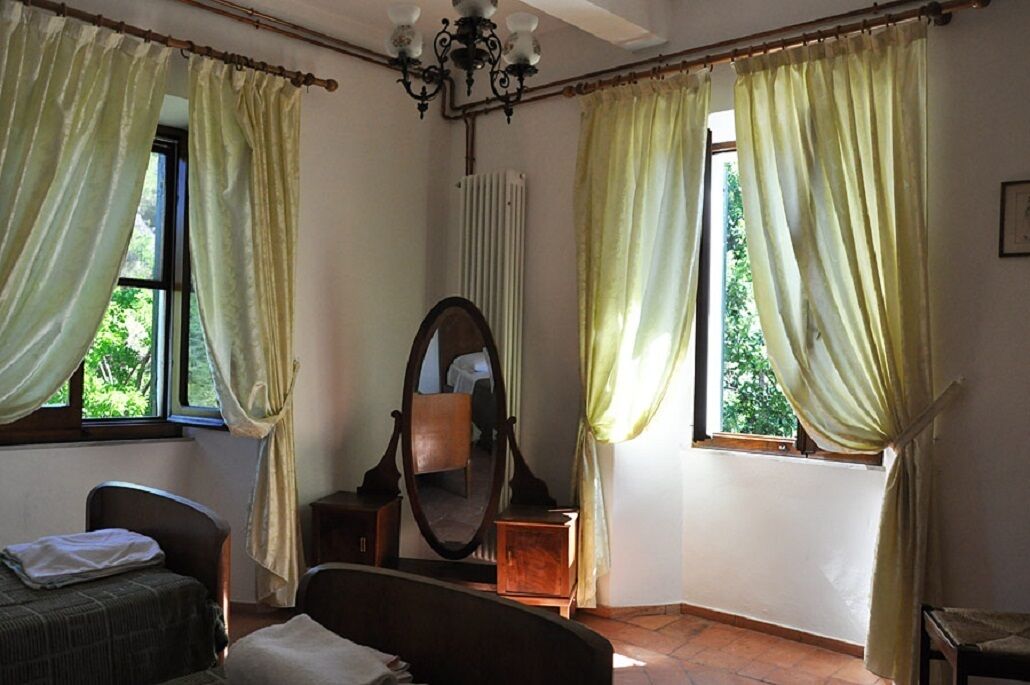 A room with a bed, a chair, a wooden cabinet, a mirror, and two windows with curtains.