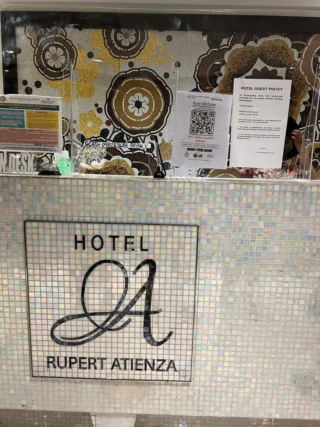 Un comptoir d’accueil d’hôtel orné d’une mosaïque, un code QR donnant accès au règlement de l’hôtel et un panneau indiquant les tarifs des chambres.