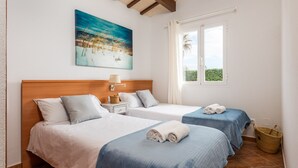 Een hotelkamer met twee bedden, een houten hoofdeinde, een schilderij van een strandlandschap, een raam met gordijnen en een rieten mand.