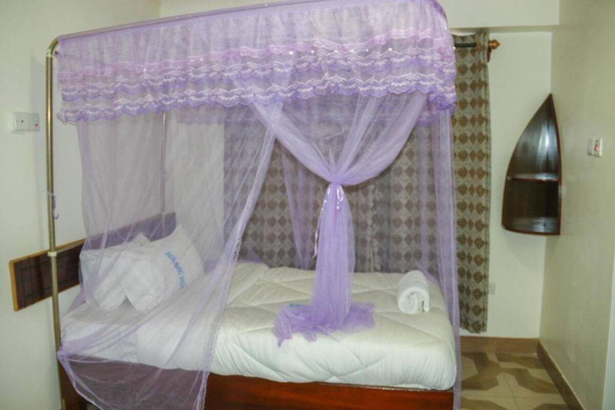 Una cama individual con dosel morado, cabecera de madera y cortina estampada al fondo.