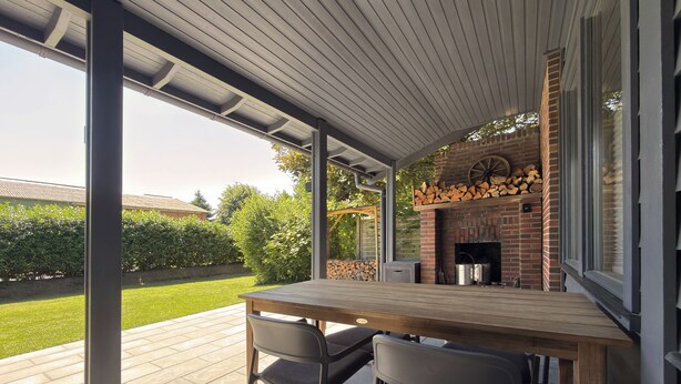 Un patio al aire libre con una mesa y sillas de madera, una chimenea de ladrillo y vistas a un jardín.