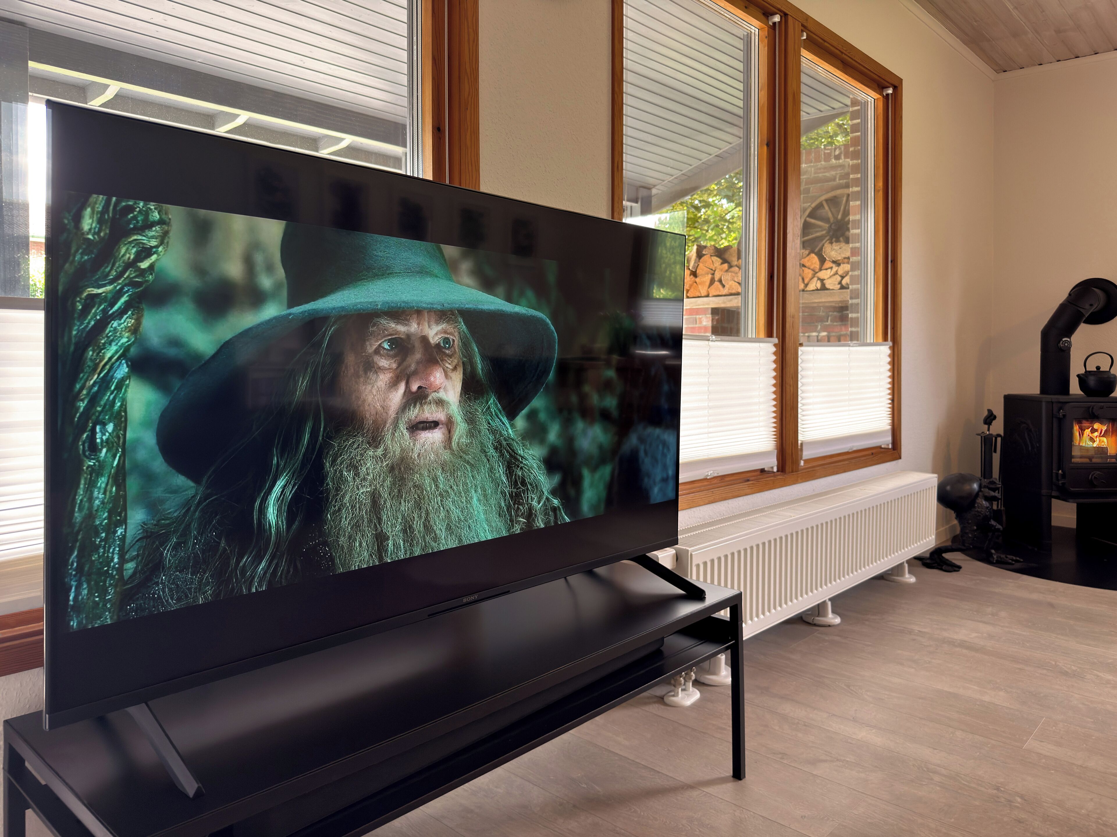 Una sala de estar moderna con un televisor de pantalla plana sobre un mueble bajo, una estufa de leña y ventanas grandes con persianas.