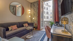 A modern living room with a sofa, a round mirror, a small side table, a kitchen area with a marble countertop, and a view of the city through large windows. - H10 Palazzo Galla (Rome)