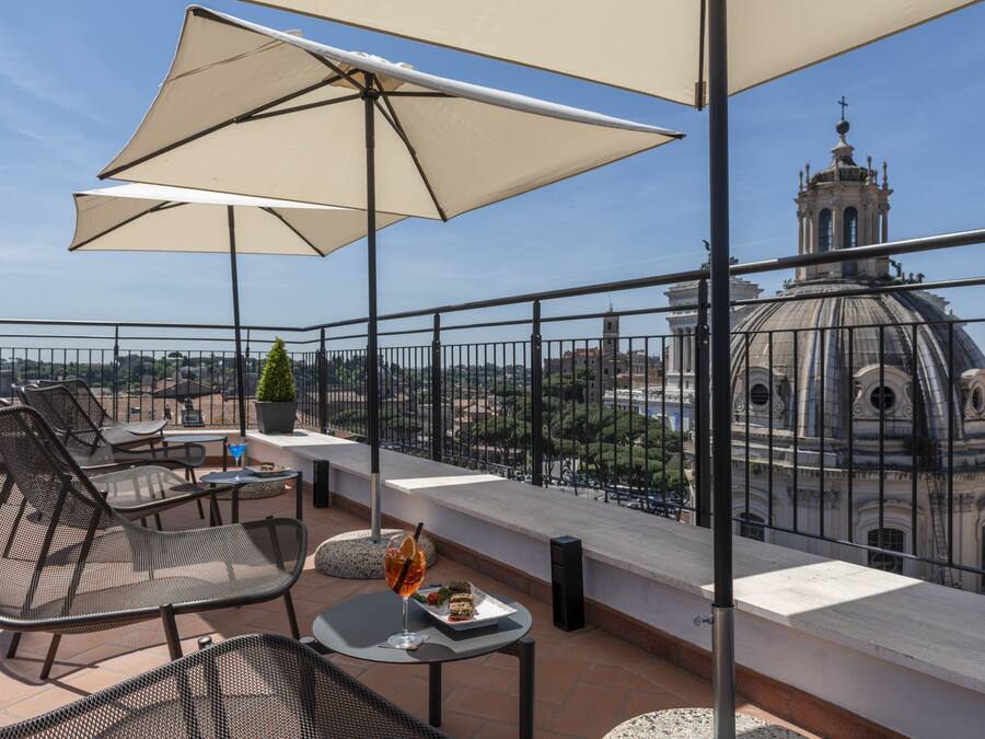 Terraço no telhado com área de estar externa, uma mesa com bebida e vista para um edifício histórico.