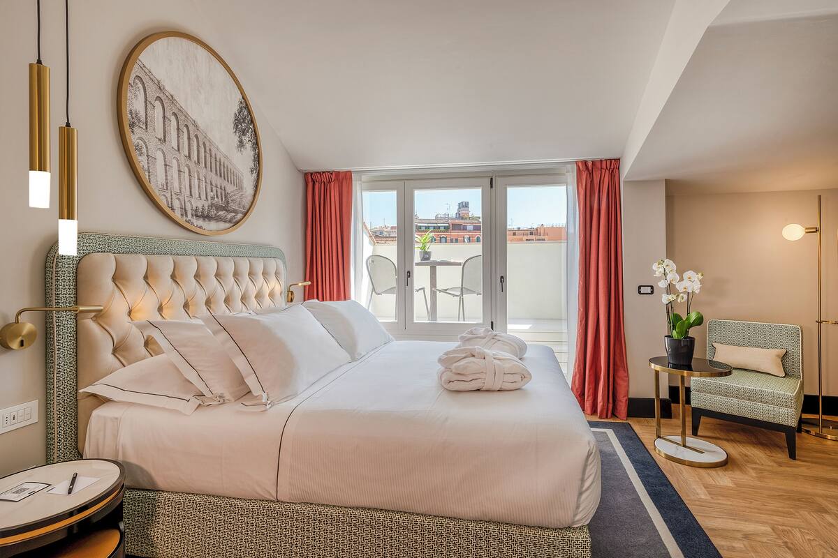 A hotel room with a large bed, a round side table, a chair, a balcony with a view of buildings, and a painting on the wall.