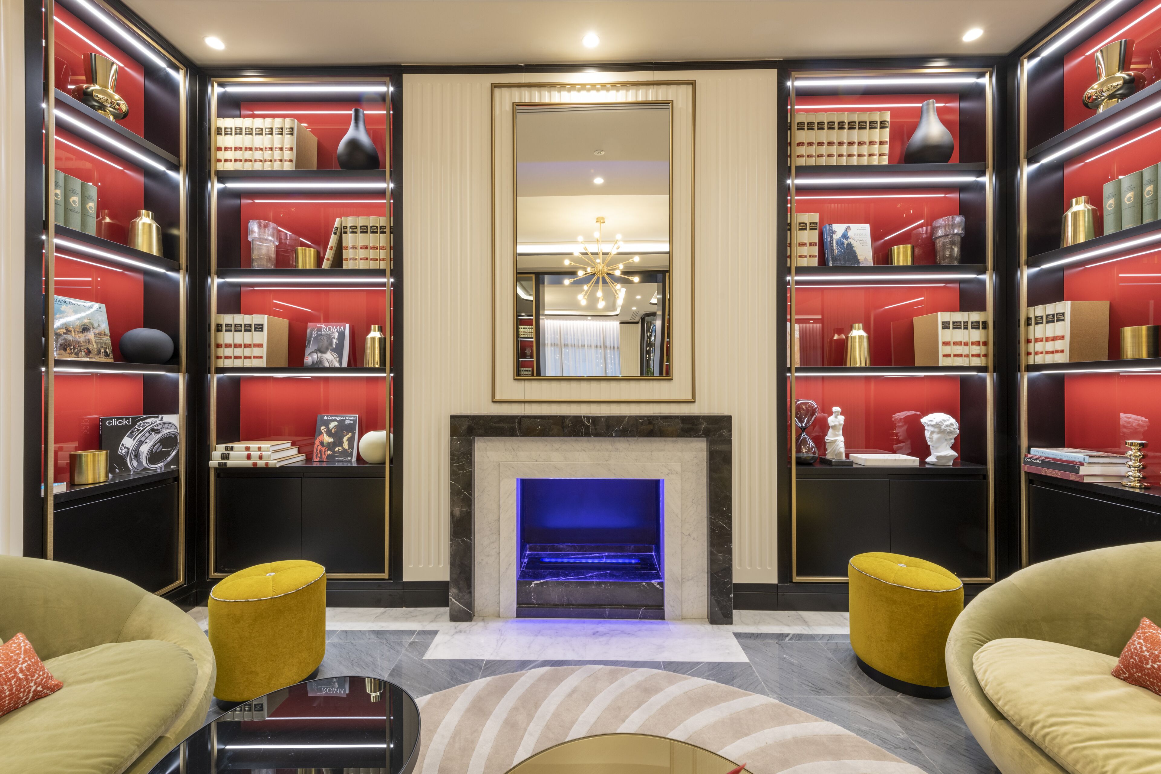 A luxurious room with a marble fireplace, red and black shelving with books and decorative items, and two yellow ottomans.