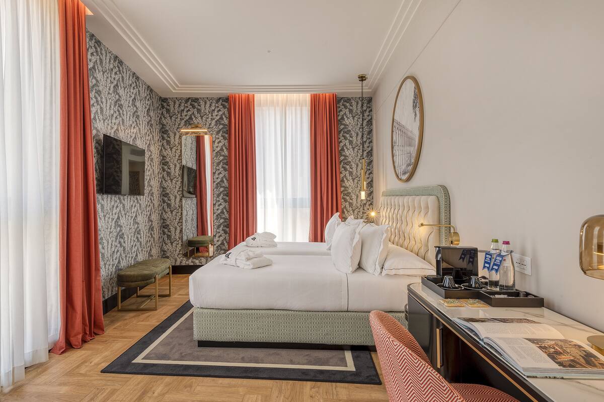 A hotel room with a large bed, a bedside table with a lamp, a bench, and a desk with a computer and papers.