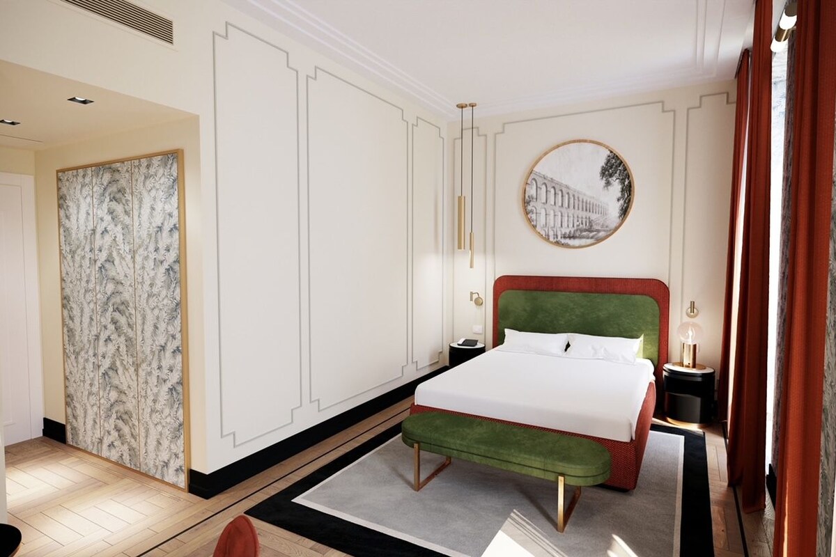 A hotel room with a large bed, a green upholstered bench, a round mirror, and a view of a building through the window.