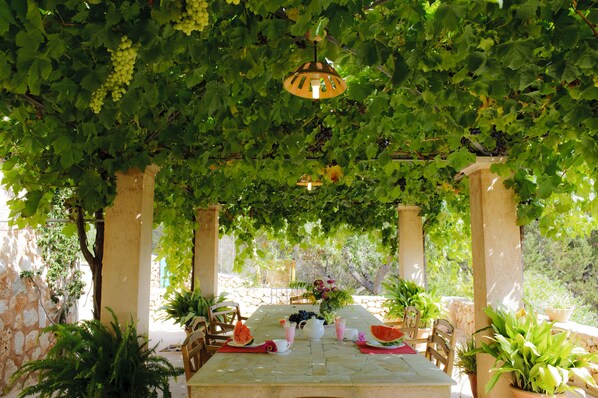 An outdoor dining area with a table set for two, surrounded by greenery and a pergola overhead.