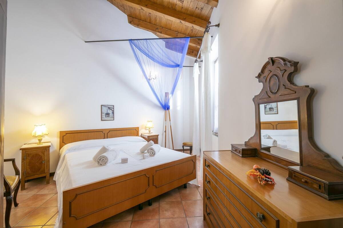 Ein Schlafzimmer mit einem hölzernen Bett, Nachttischlampen, einer Kommode, einem Spiegel und einem Fenster mit blauem Vorhang.