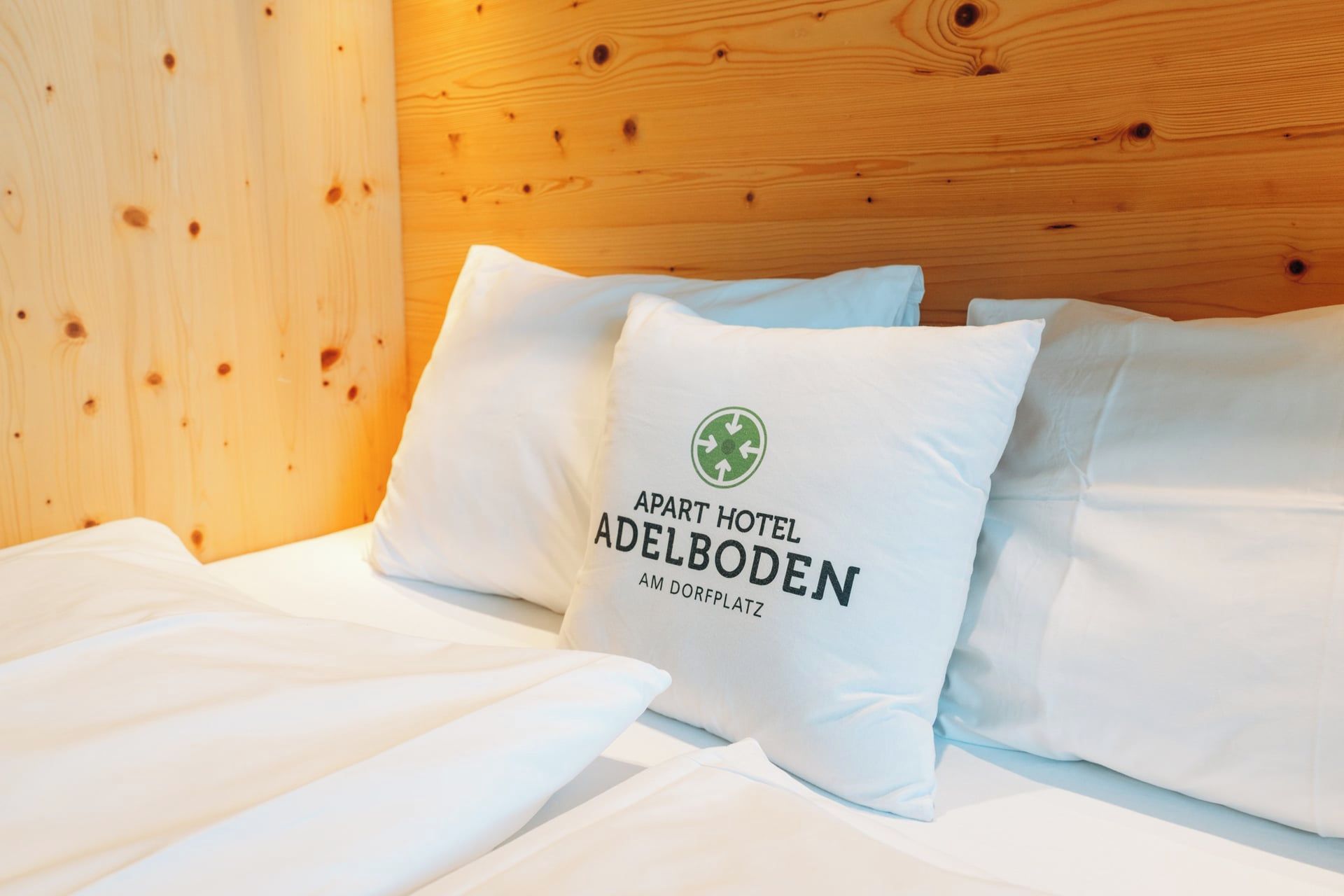 Una cama con ropa de cama y almohadas blancas, una de ellas con un logotipo y texto que indica que pertenece al Apart Hotel Adelboden.