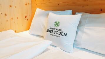 Una cama con ropa de cama y almohadas blancas, una de ellas con un logotipo y texto que indica que pertenece al Apart Hotel Adelboden.