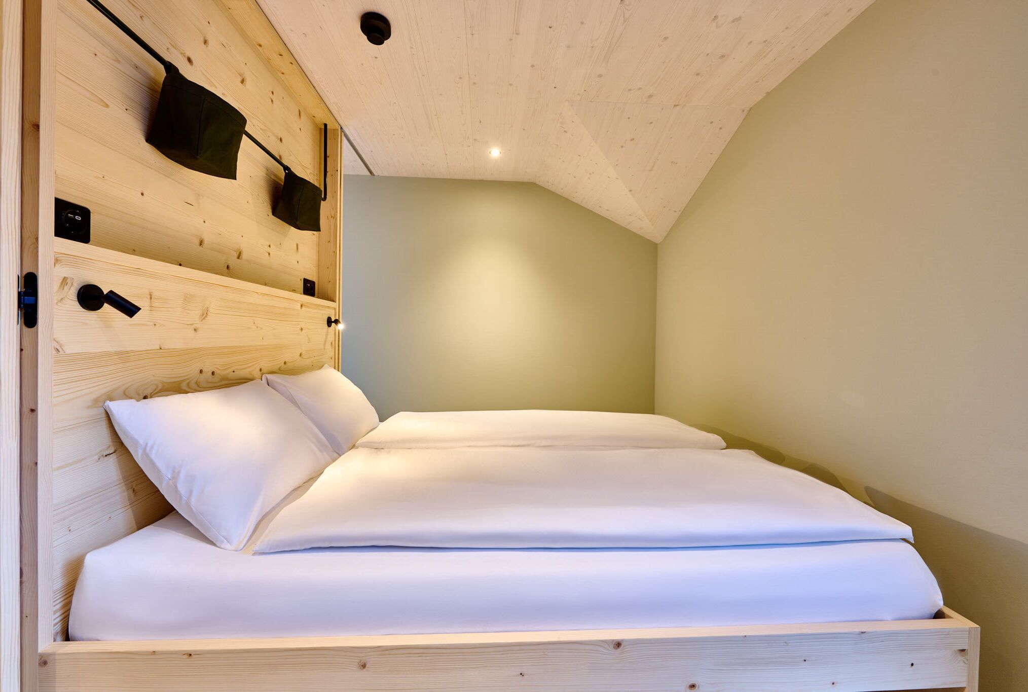 A wooden bunk bed with white bedding, a headboard with storage pockets, and a sloped ceiling.