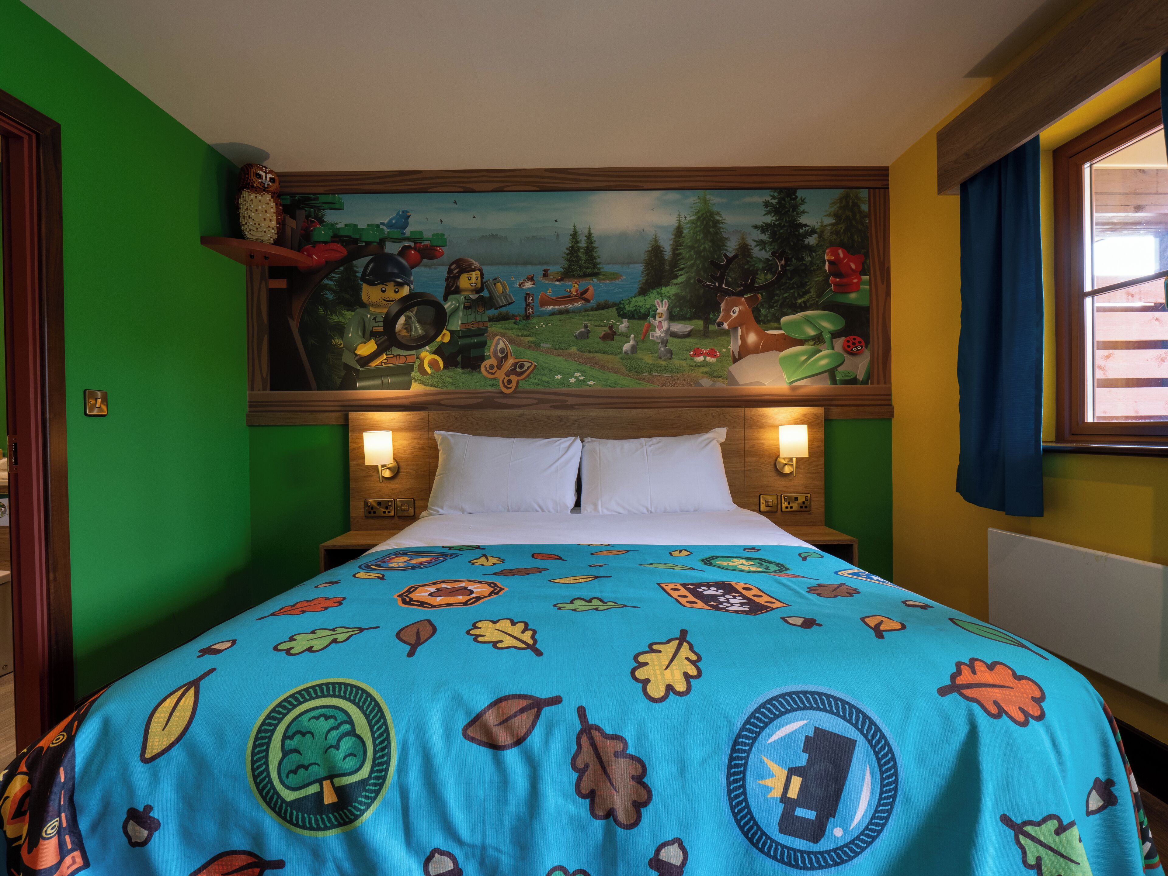 A hotel room with a bed featuring a colorful leaf-patterned bedspread, a mural depicting a forest scene on the wall, and a window with blue curtains.