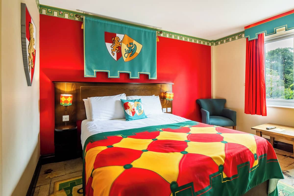 A hotel room with a bed, a chair, a window with curtains, and a wall with a decorative shield emblem.