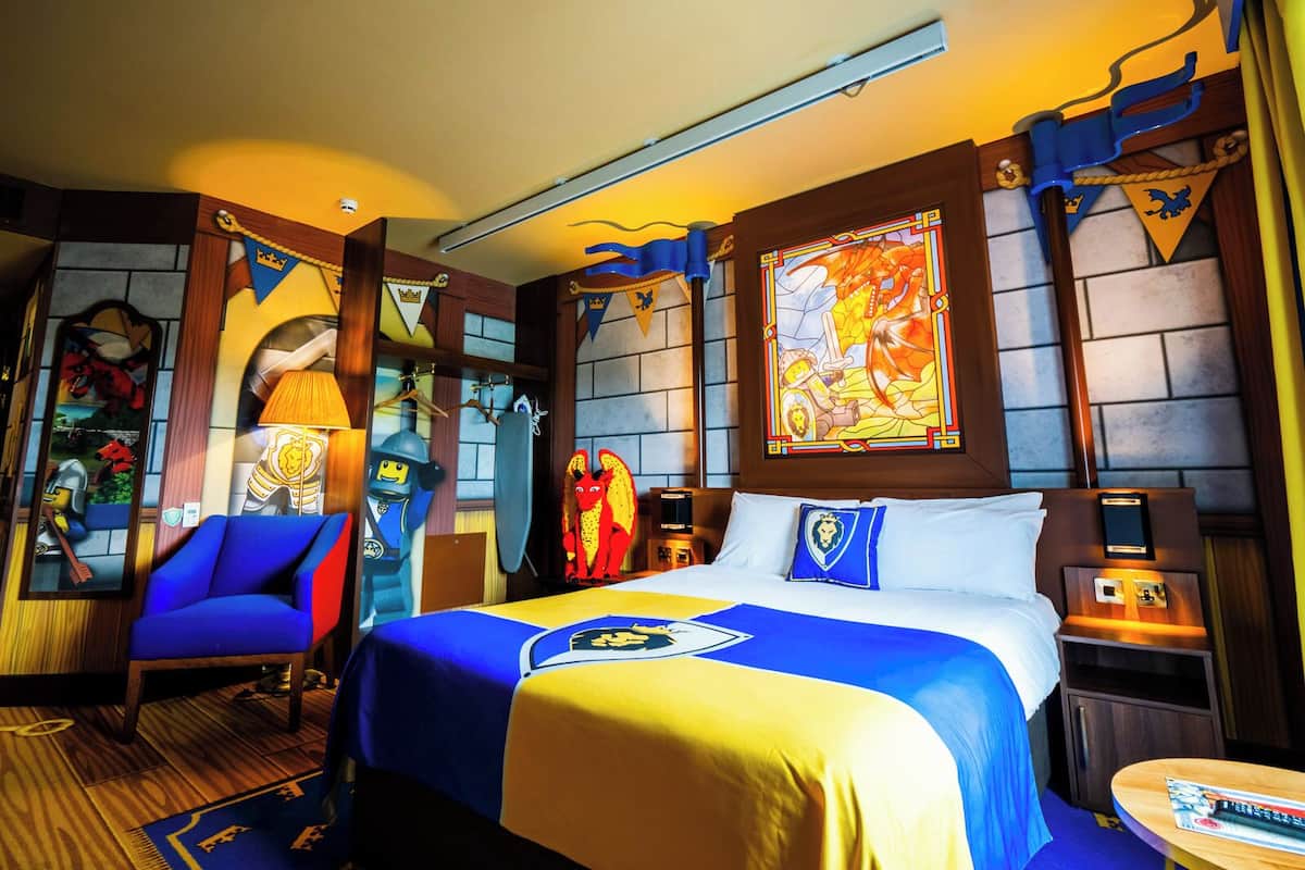 A hotel room with a bed, a chair, a nightstand, and a wall with a stained glass window.