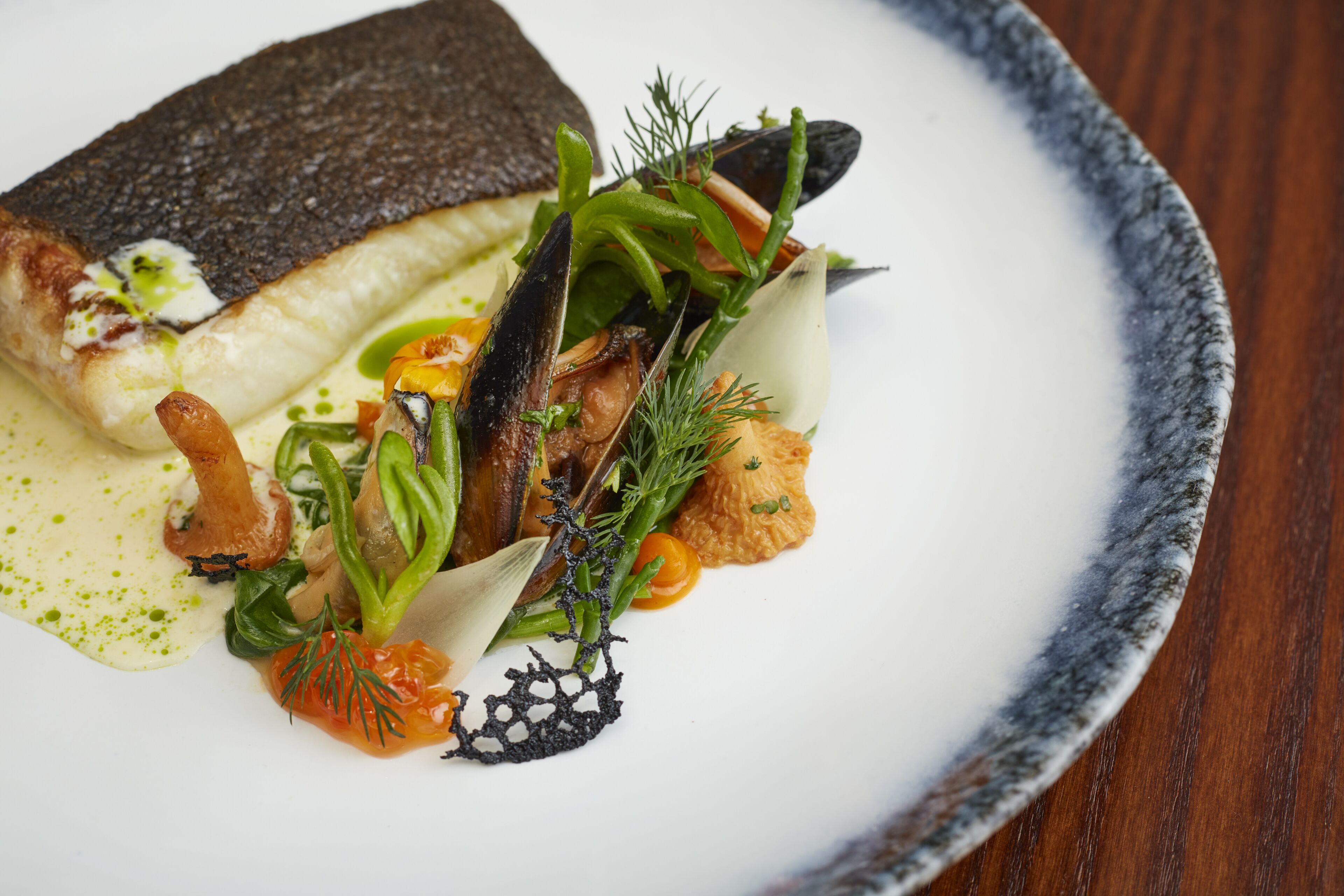 A plate of food with a fish, mussels, greens, and a sauce.
