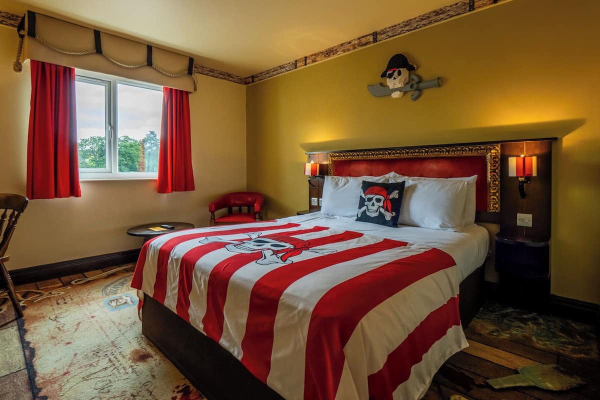 A hotel room with a large bed, red curtains, a small chair, a nightstand, and a window with a view of trees.