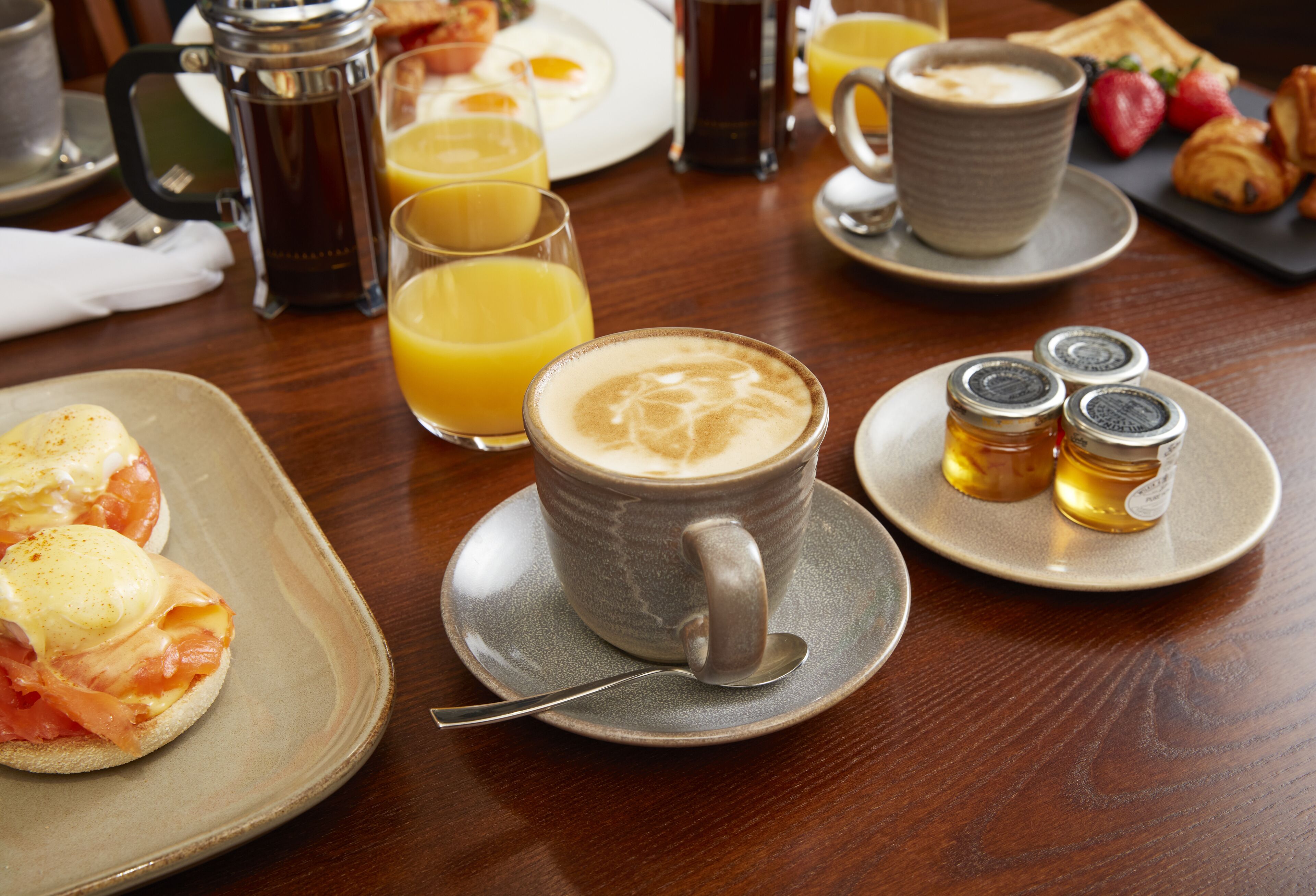 Un desayuno con huevos Benedict, zumo de naranja, café, croissants y salsas en una mesa de madera.