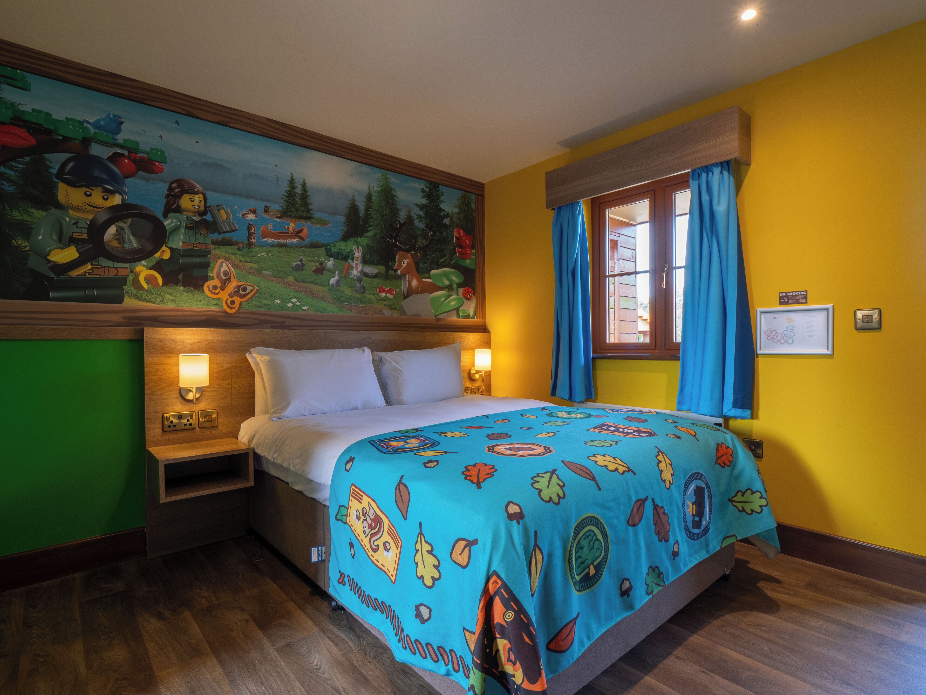 A bedroom with a bed, a nightstand, a wall mural, a window with curtains, and a 'No Smoking' sign.