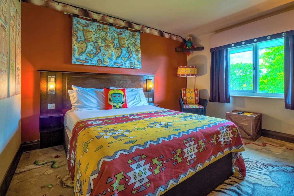 A hotel room with a bed, a nightstand, a lamp, a chair, and a window with a view of greenery.