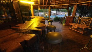 A rustic bar area with wooden stools and a bar counter, surrounded by a wooden deck and a pool view. - Naiberi Resort (Eldoret)