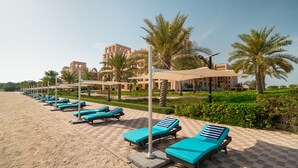 Ein Liegebereich am Strand mit Liegestühlen und Sonnenschirmen, Palmen und einem Gebäude im Hintergrund.