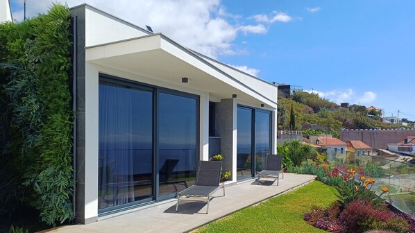 Una casa moderna con amplios ventanales correderos, un patio con mobiliario exterior y un jardín bien cuidado.
