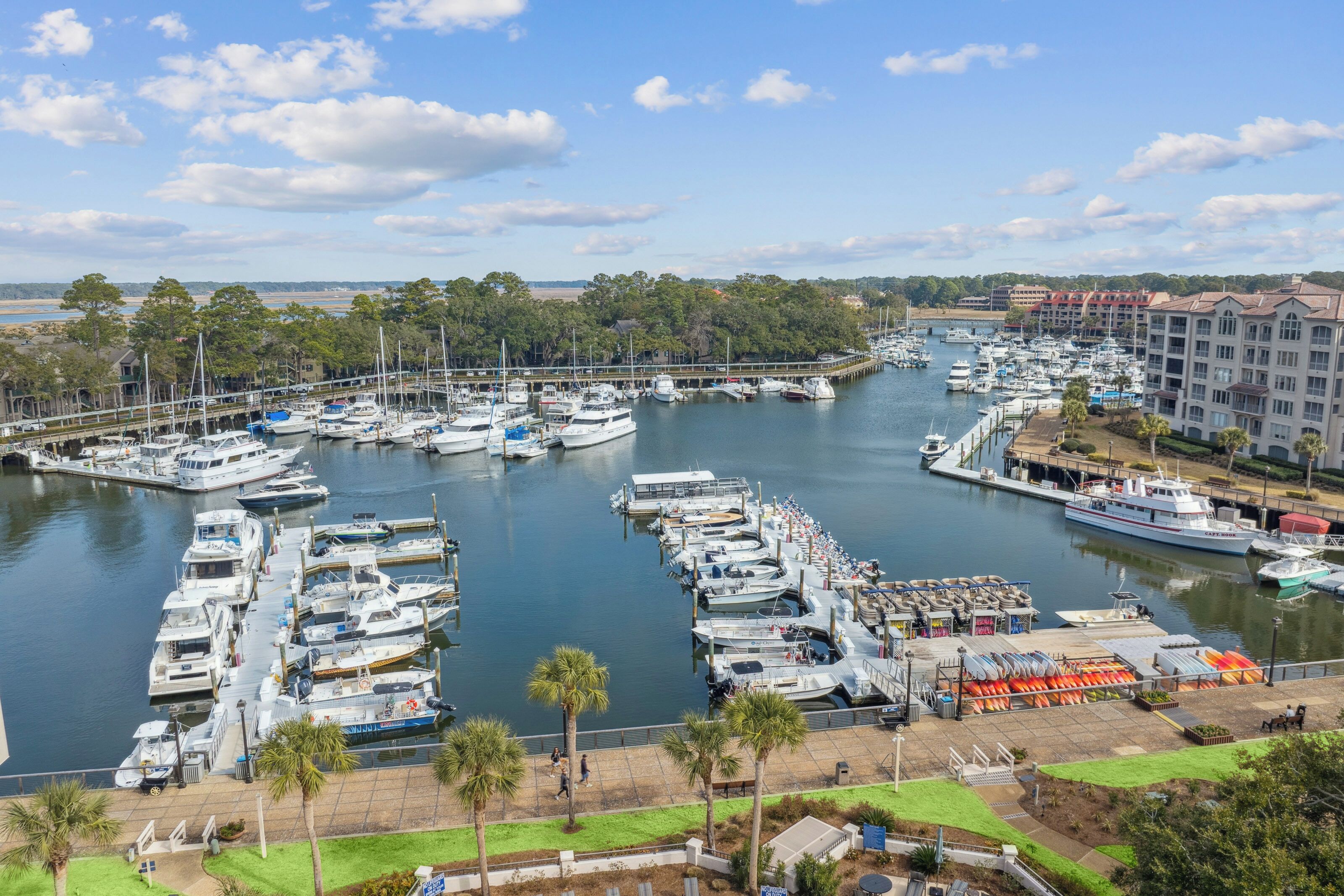 A marina with numerous boats docked, a pedestrian walkway, and a building with a red roof in the background.