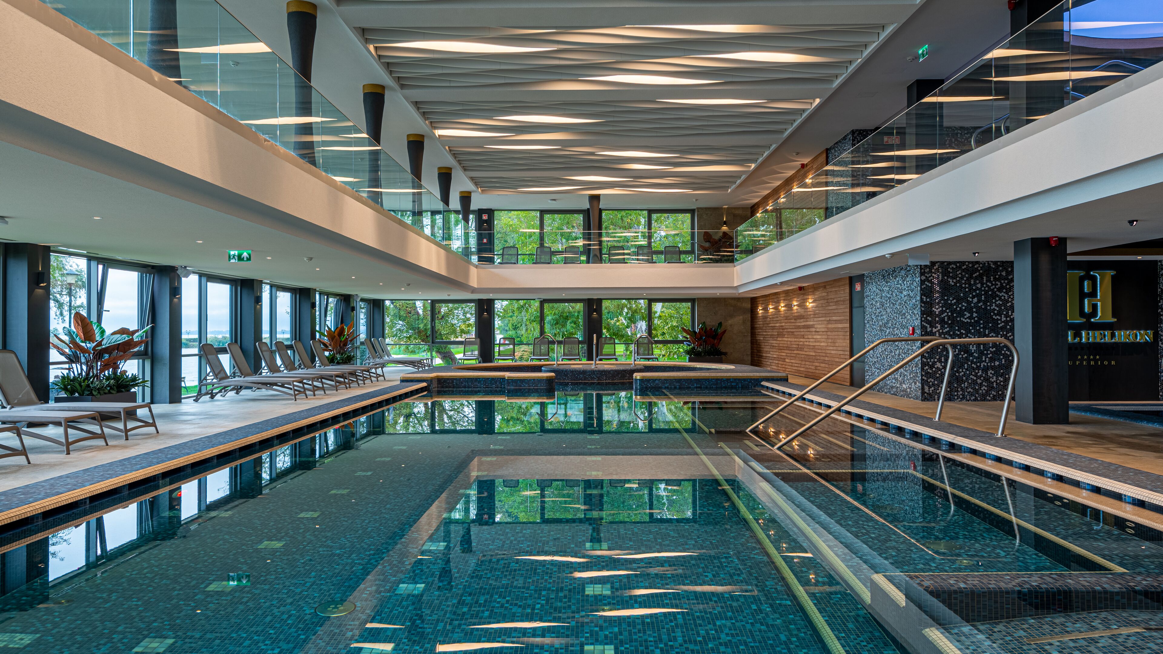 Una piscina interior moderna con paredes de vidrio transparente, sillones de descanso y vistas a zonas verdes.