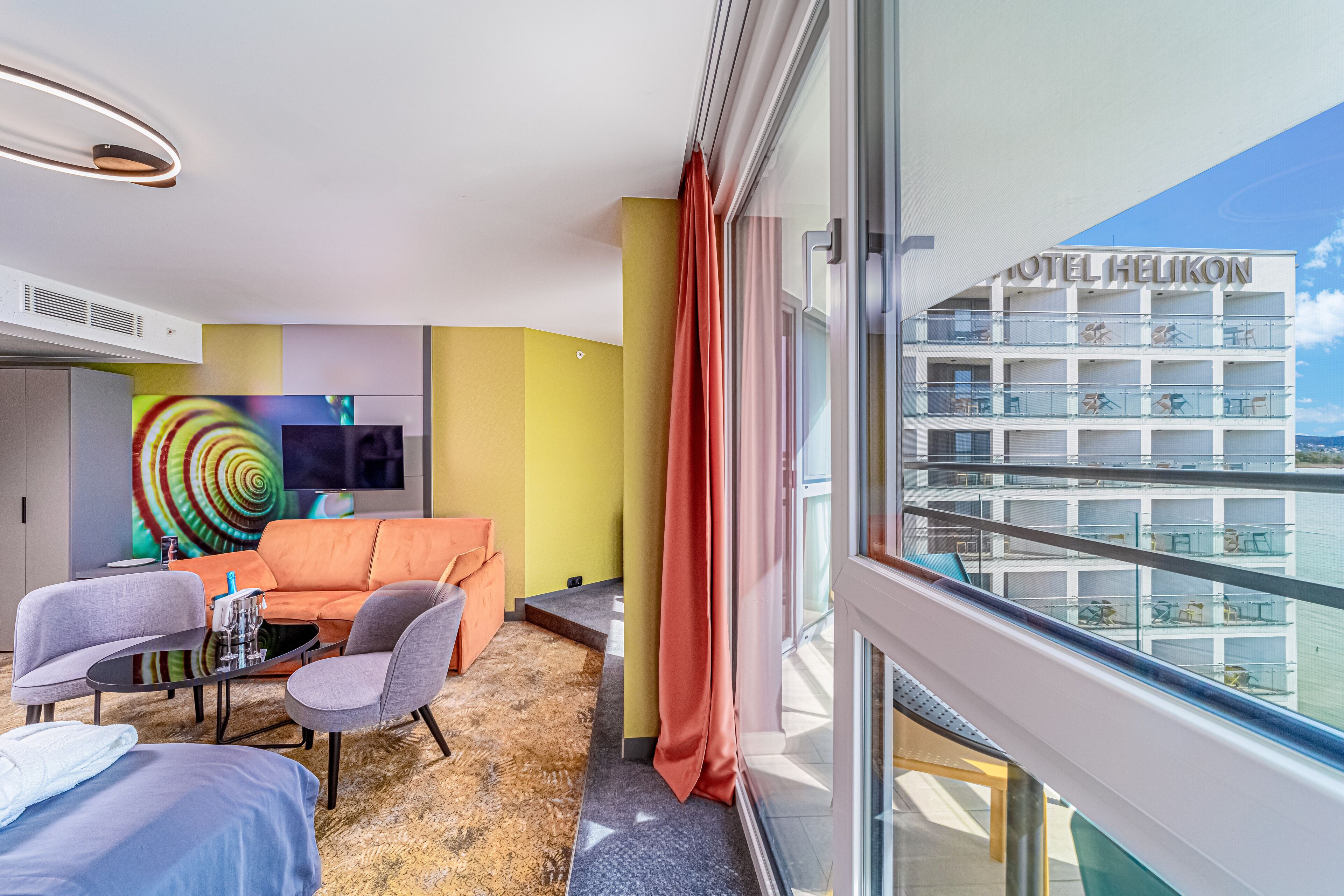 A modern hotel room with a balcony, a large TV, and a colorful abstract painting.