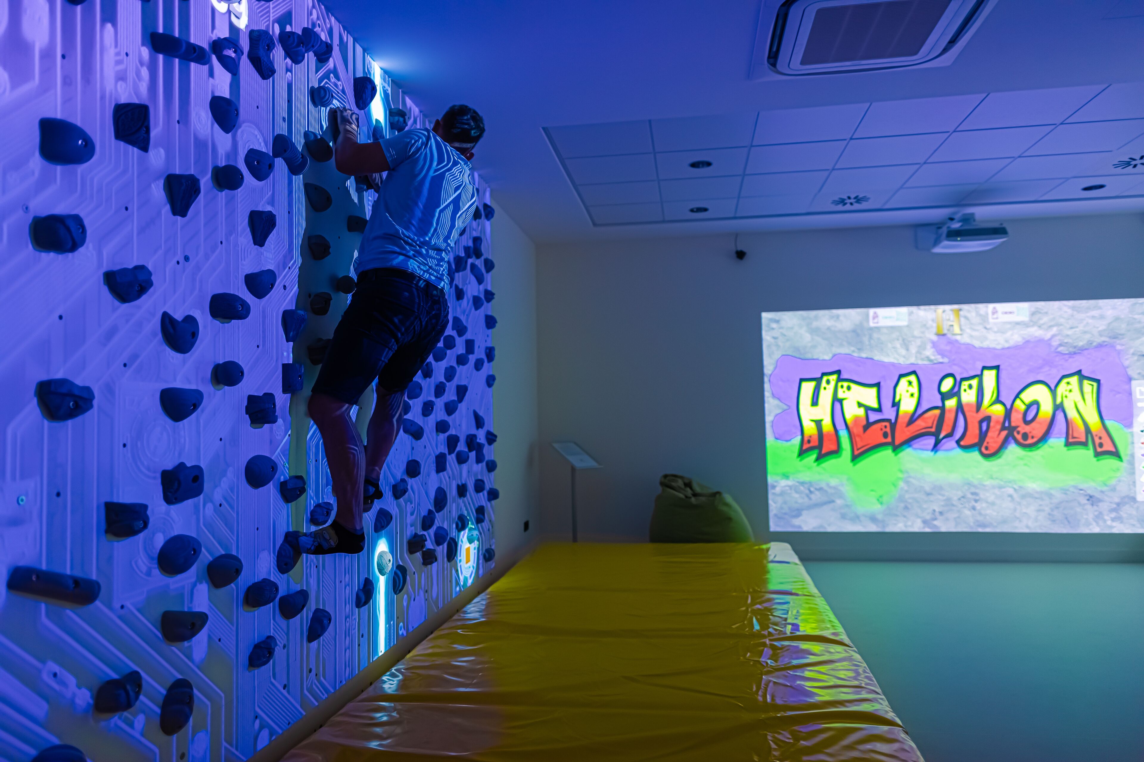 Una pared de escalada con una pantalla de proyección que muestra 'HELION' al fondo.