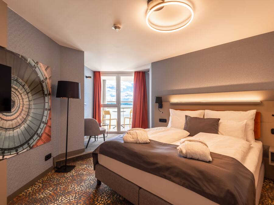 A hotel room with a large bed, a flat-screen TV mounted on the wall, a floor lamp, and a view of the outside balcony.