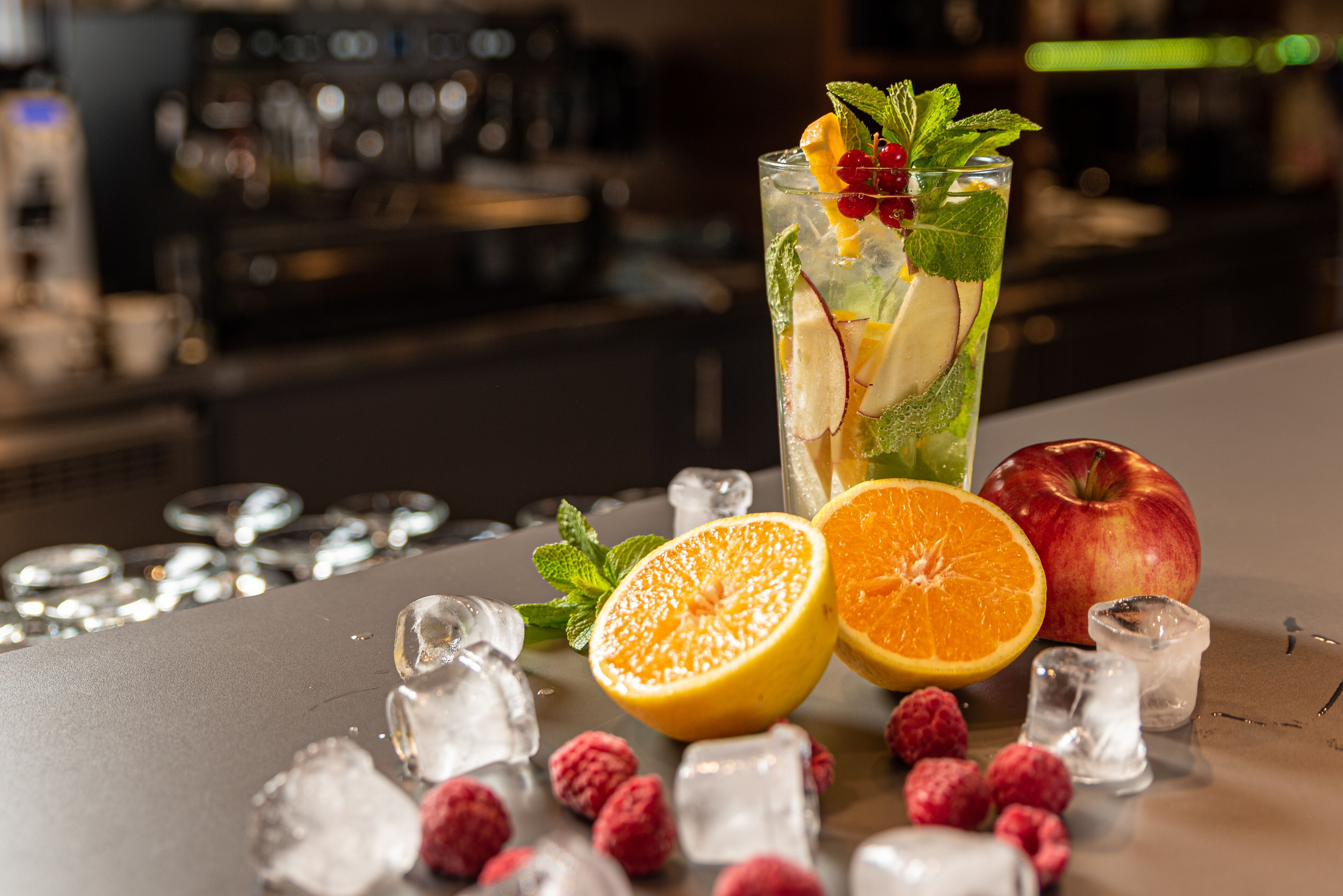 Eine Bar mit einem hohen Glas, das einen Cocktail enthält, garniert mit Früchten und Minze, umgeben von Eiswürfeln, einer halben Orange, einem ganzen Apfel und Himbeeren.