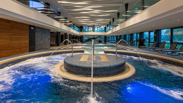 Una piscina interior con un jacuzzi central, barandillas de vidrio y sillones de descanso.